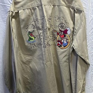May include: A beige long-sleeve button-down shirt with an embroidered map of an island, a crocodile, and Captain Hook on the back.