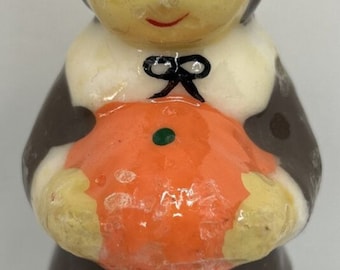 May include: A vintage candle shaped like a pilgrim figure, holding an orange pumpkin. The candle has a brown hat with an orange band, a white collar, and a black bow. The figure's face is yellow with black eyes and a red smile.
