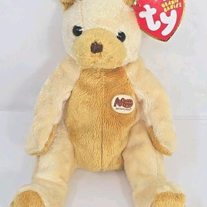 Peut inclure: Un ours en peluche beige et crème, avec un nez marron et des yeux noirs. Il porte un écusson Bass Pro Shops Old Country Store et une étiquette rouge en forme de cœur Ty Beanie Babies.