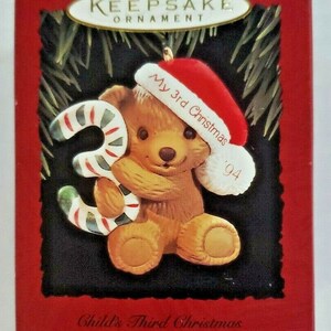 May include: A brown teddy bear Christmas ornament wearing a red Santa hat with the text "My 3rd Christmas '94". The bear holds a candy cane-striped number "3". The ornament is from the Child's Age Collection.