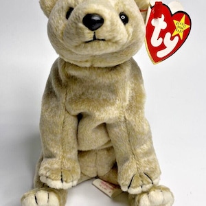 May include: A tan plush bear toy with a black nose and eyes. The bear has a red heart-shaped tag attached to its ear. The tag reads "Beanie Original Baby" and features the "ty" logo.
