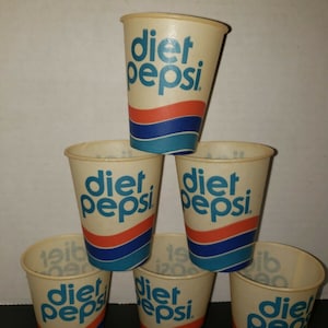 May include: A stack of six vintage Diet Pepsi paper cups. The cups are beige with the words "diet pepsi" in blue, and a red and blue stripe design. The cups are arranged in a pyramid shape.