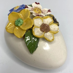 May include: A white ceramic egg with a floral design. The egg is decorated with yellow, white, and blue flowers, and green leaves.