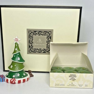 May include: A set of 12 tealight candles in a box with a green Christmas tree design. The box has the text "VD455205 Universal Tealight Spruce in the Snow" and "12 Tealight Candles"