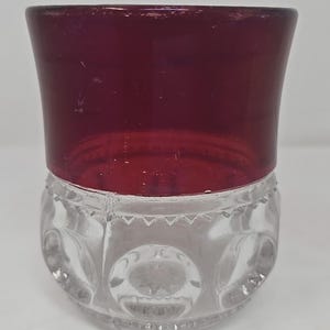 May include: A two-part glass container with a deep red top and a clear, patterned base. The top is a solid, smooth color, while the base features a repeating circular design. The container is likely for storage or display.