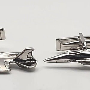 May include: A pair of silver cufflinks shaped like fighter jets. The cufflinks are detailed and have a realistic design.