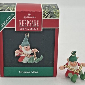 May include: Miniature Hallmark Keepsake ornament of an elf stringing beads. The elf wears a red coat, green hat and shoes, and a white beard. The ornament is next to its original box, which reads "Stringing Along."