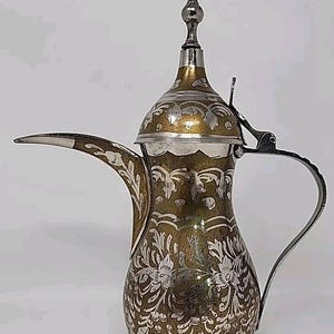 May include: A silver and gold Arabic coffee pot with intricate floral designs. The pot has a spout and a handle, and a decorative lid with a finial.
