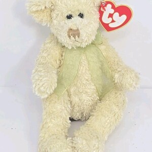 May include: A cream-colored plush teddy bear with a green vest and a red heart-shaped tag. The bear has black eyes and a brown nose. The vest is trimmed with gold thread. The bear is a stuffed animal.