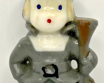 May include: A vintage figurine of a person in a gray outfit and hat, holding a brown object. The figurine has a white face with blue eyes and a red nose. The figure is approximately 4 inches tall.