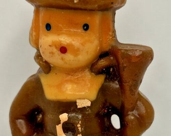 May include: A vintage candle shaped like a pilgrim figure. The candle is brown with a tan face, blue eyes, and a red mouth. The pilgrim wears a hat and holds a staff. A decorative item for Thanksgiving or fall.