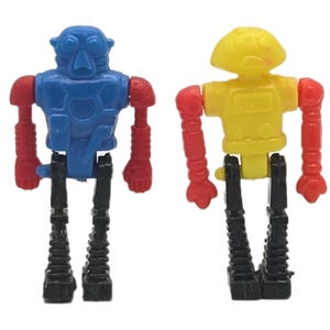 May include: Two small, plastic robot toys. One is blue with red arms and the other is yellow with red arms. Both have black legs and are standing upright. The robots have a retro, vintage aesthetic.