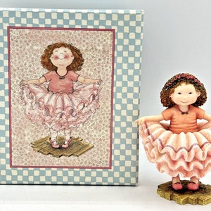 May include: A figurine of a girl wearing a pink tutu and a pink shirt. She is standing on a wooden platform and has a happy expression on her face. The figurine is packaged in a blue and white checkered box.