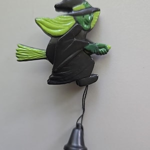 May include: A Halloween-themed decoration featuring a witch riding a broom. The witch is depicted in black attire with a green face and hat, holding a green broom. A small bell hangs below the witch, attached by a string. The decoration is likely made of plastic.