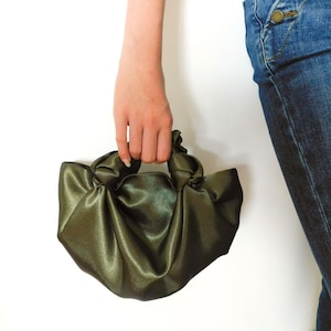 Pu&ograve; includere: Una borsa a mano in satin verde con un design annodato. La borsa &egrave; tenuta da una mano.