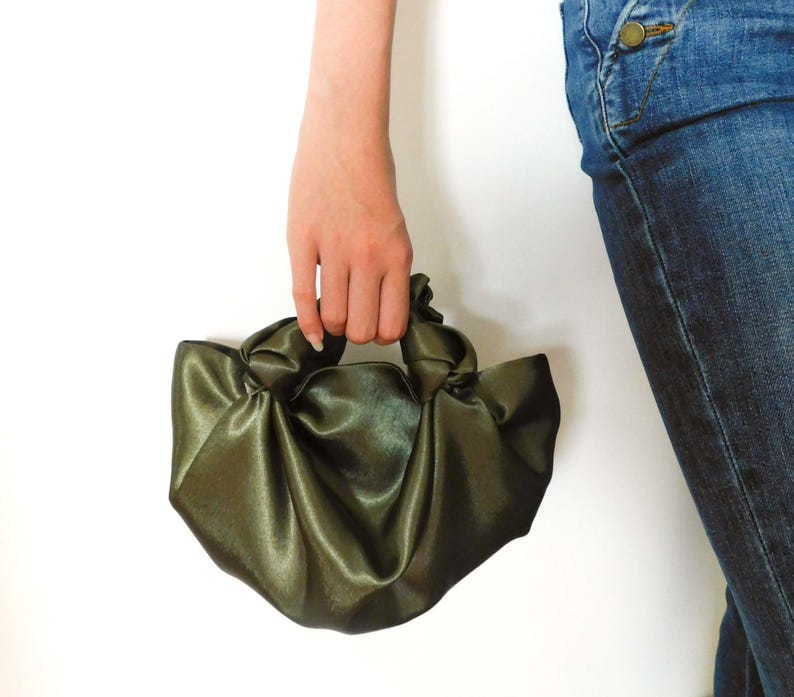 Pu&ograve; includere: Una borsa a mano in satin verde con un design annodato. La borsa &egrave; tenuta da una mano.