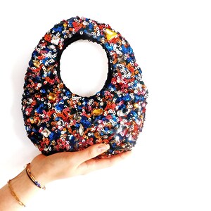 以下が含まれることがあります： カラフルなスパンコールで覆われた、小さな楕円形のハンドバッグ。バッグは黒い内側と円形のハンドル開口部が特徴です。スパンコールは赤、青、オレンジ、金の色合いで、きらめく効果を生み出しています。バッグは白い背景に対して保持されています。