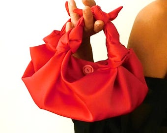 Puede incluir: Un bolso de mano de estilo cartera de color rojo brillante con un cierre de botón único. El bolso está hecho de una tela suave y lisa y tiene un asa anudada.