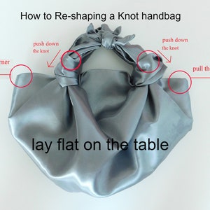 May include: A gray satin knot handbag laid flat on a white surface. The image shows how to reshape the bag by pulling the corners and pushing down on the knot.
