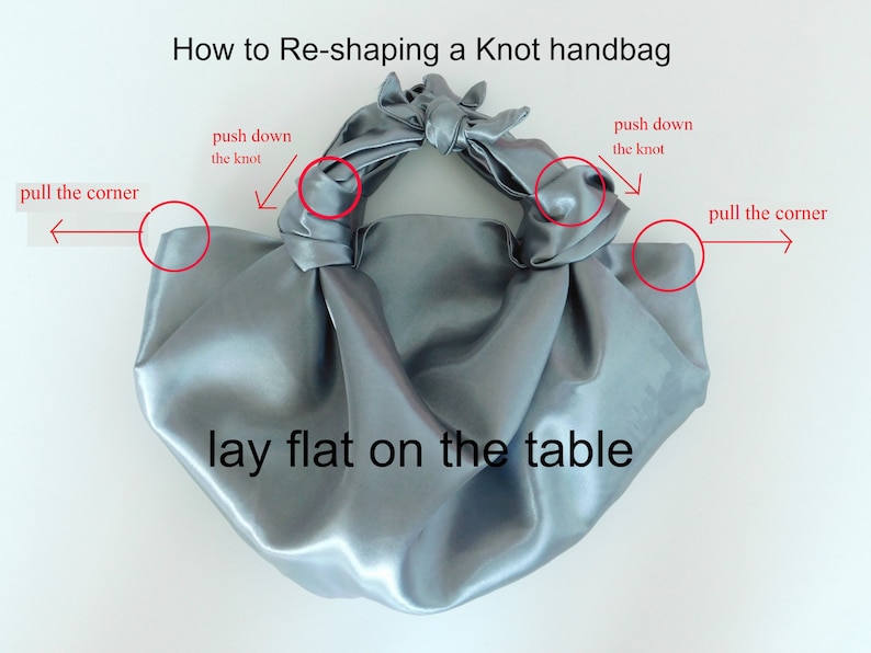 May include: A gray satin knot handbag laid flat on a white surface. The image shows how to reshape the bag by pulling the corners and pushing down on the knot.