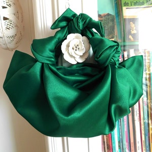 May include: A green satin handbag with a large white flower brooch. The bag is gathered and knotted at the top, creating a unique and stylish design.
