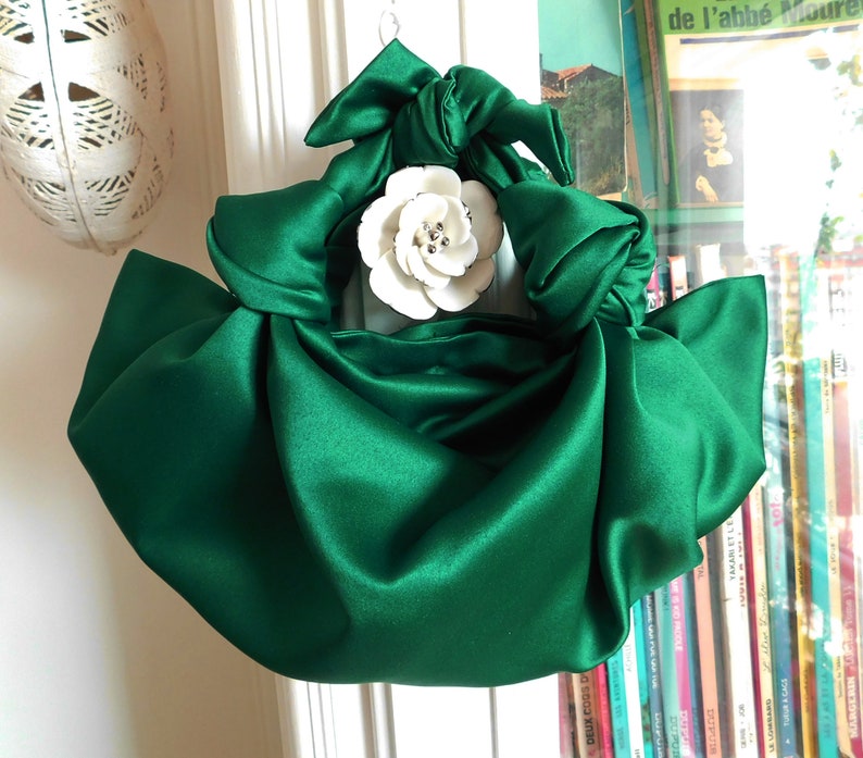 May include: A green satin handbag with a large white flower brooch. The bag is gathered and knotted at the top, creating a unique and stylish design.