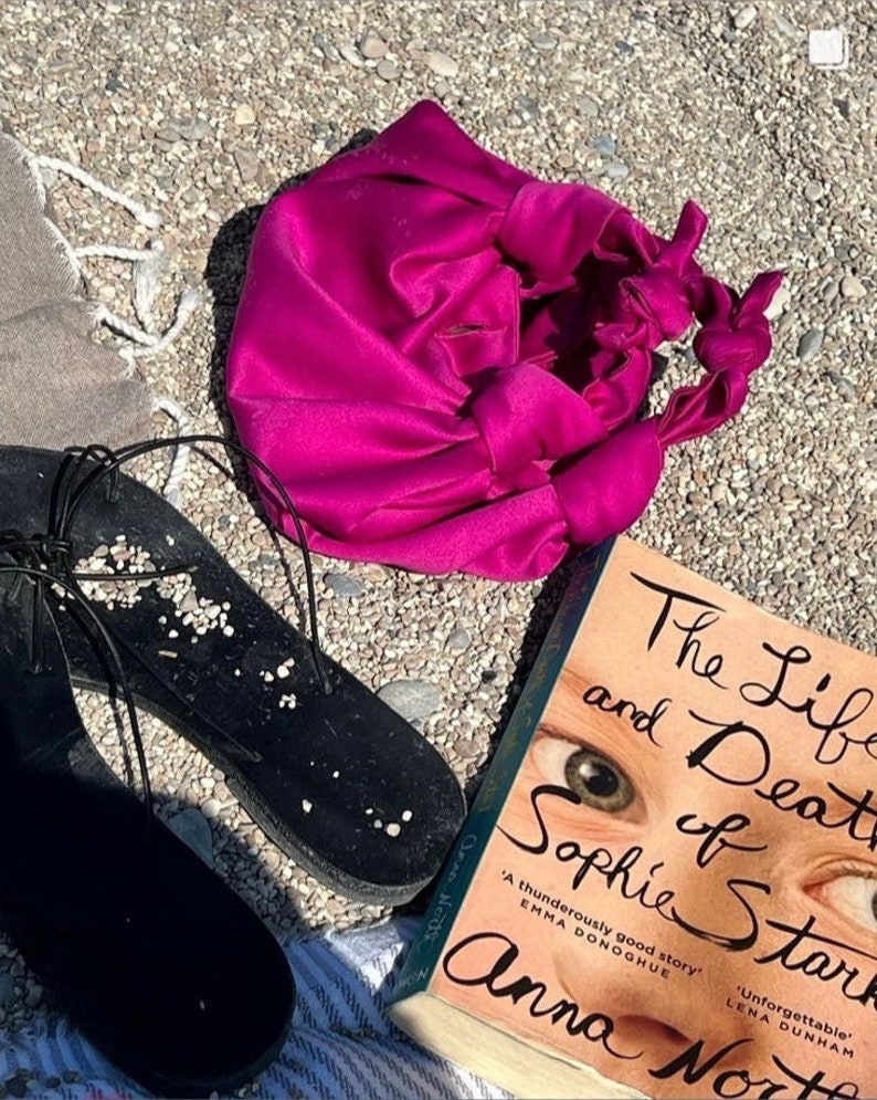 Może przedstawiać: Książka w miękkiej oprawie zatytułowana "The Life and Death of Sophie Stark" autorstwa Anny North. Okładka książki przedstawia zbliżenie oczu młodej dziewczyny. Książka leży na plaży z r&oacute;żową torbą i czarnymi sandałami.