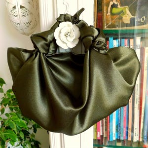 Pu&ograve; includere: Una borsa a mano in satin verde con un dettaglio floreale bianco. La borsa &egrave; raccolta e annodata nella parte superiore, creando un design unico ed elegante.