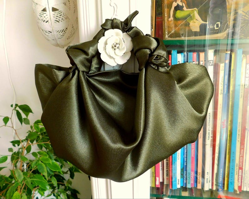 Pu&ograve; includere: Una borsa a mano in satin verde con un dettaglio floreale bianco. La borsa &egrave; raccolta e annodata nella parte superiore, creando un design unico ed elegante.
