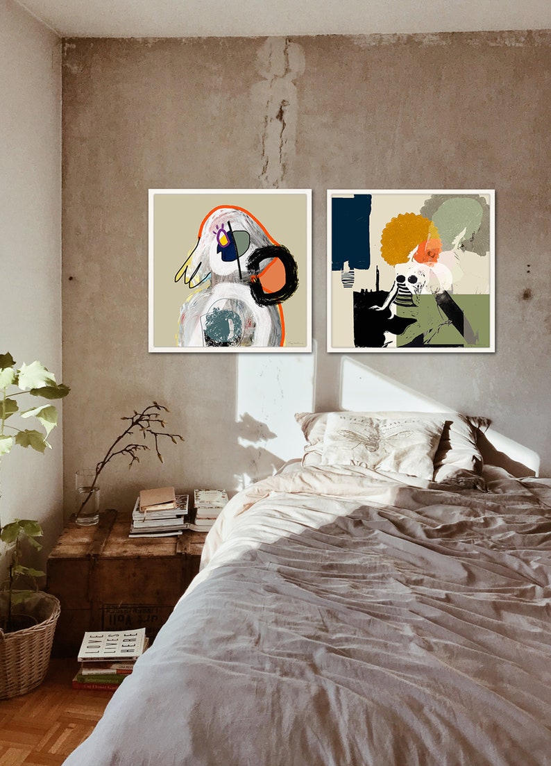 May include: Two framed abstract paintings with colourful geometric shapes and figures hang on a light brown wall above a bed with a white pillow and a beige blanket.