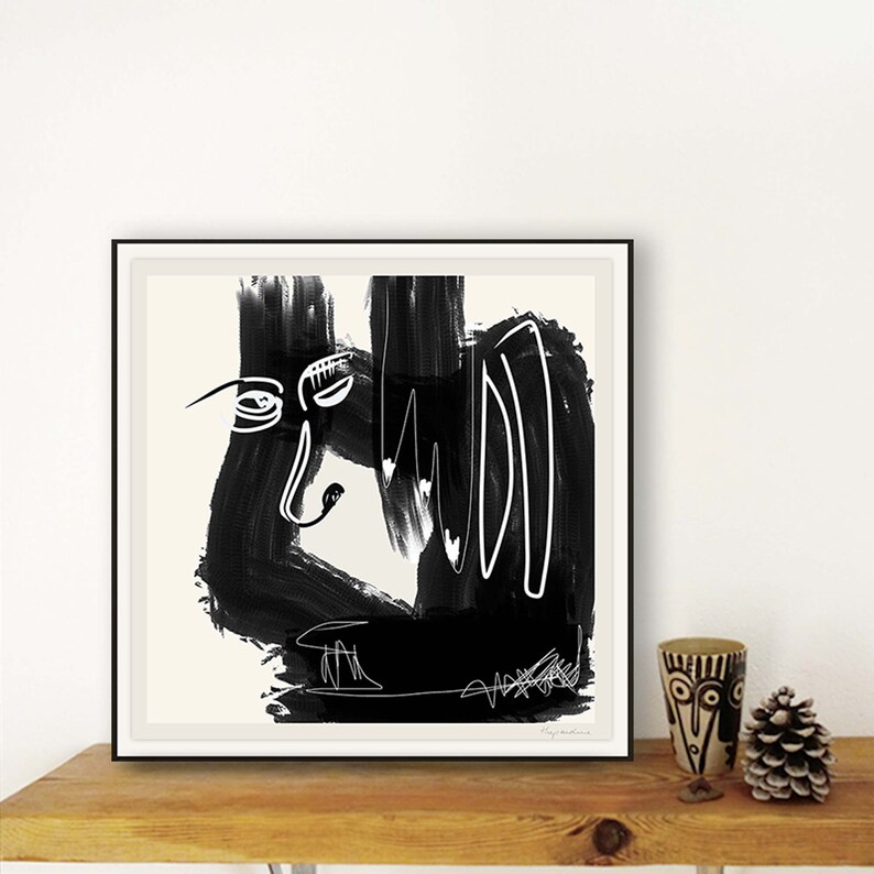May include: Black and white abstract art print with a stylized face and bold brushstrokes. The print is framed in a black frame and is on a wooden shelf.