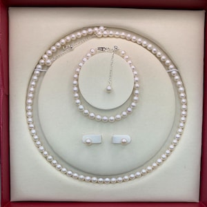 May include: A pearl jewelry set displayed in a red box. The set includes a necklace, bracelet, and a pair of stud earrings. The pearls are white and round, and the necklace and bracelet have a silver clasp. The set is presented on a cream-colored surface.