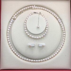 May include: A pearl jewelry set displayed in a red box. The set includes a necklace, bracelet, and earrings. The necklace and bracelet are composed of small, round pearls. The earrings are pearl studs. The jewelry is presented on a white, cushioned surface.