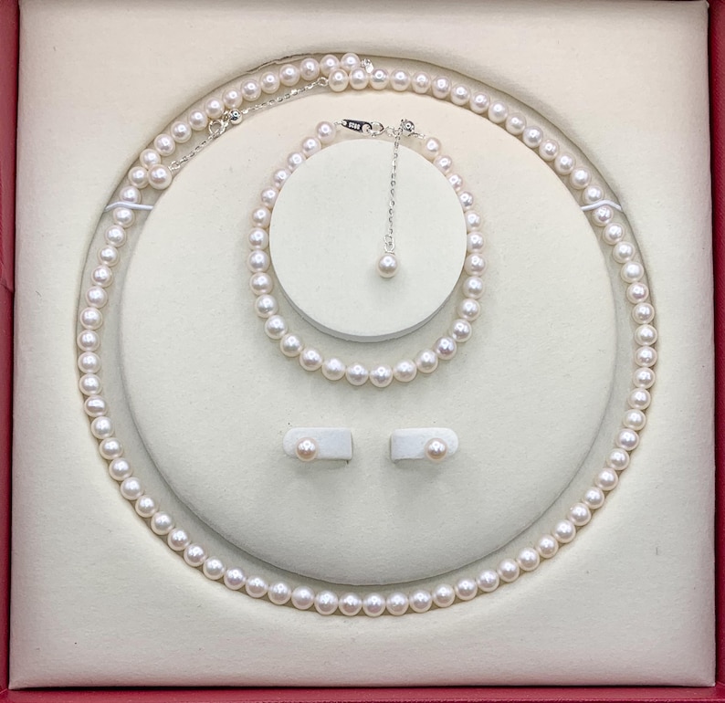 May include: A pearl jewelry set displayed in a red box. The set includes a necklace, bracelet, and earrings. The necklace and bracelet are composed of small, round pearls. The earrings are pearl studs. The jewelry is presented on a white, cushioned surface.