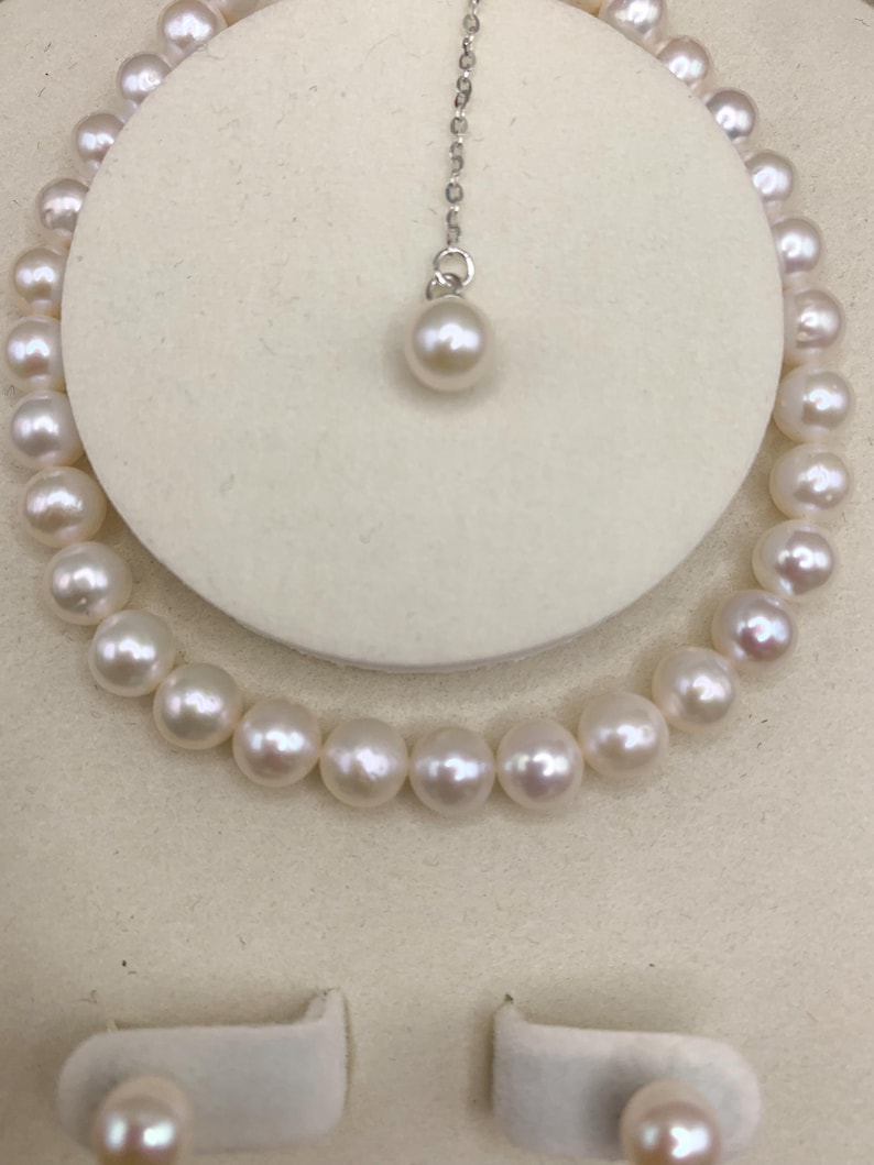 May include: A pearl necklace and matching earrings set. The necklace is a strand of round, white pearls. A single pearl pendant hangs from a delicate silver chain. The earrings are also pearl studs. The jewelry is displayed on a white background.