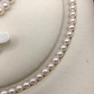 May include: A close-up view of a delicate pearl necklace. The necklace features a strand of round, lustrous pearls, each with a creamy white color. The pearls are strung together, creating a graceful curve. The background is a soft, off-white fabric.