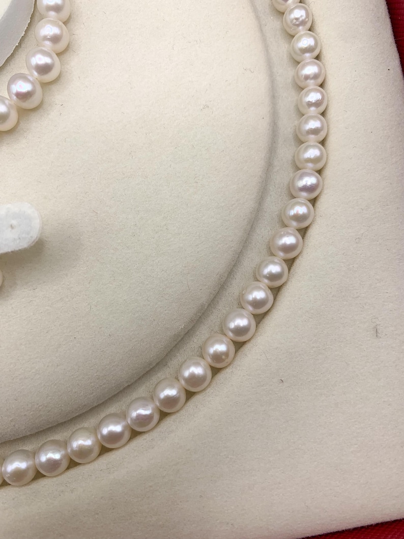 May include: A close-up view of a delicate pearl necklace. The necklace features a strand of round, lustrous pearls, each with a creamy white color. The pearls are strung together, creating a graceful curve. The background is a soft, off-white fabric.