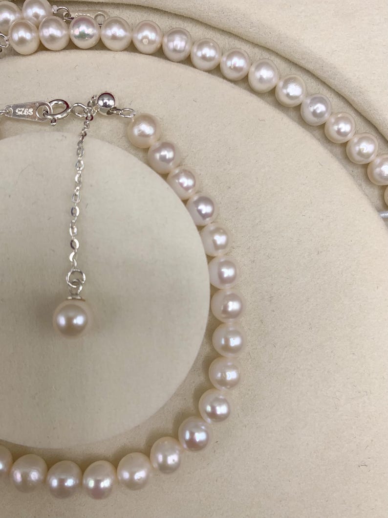 May include: A delicate pearl necklace and bracelet set. The jewelry features lustrous, off-white pearls strung together with a silver chain and clasp. The bracelet is partially visible, showcasing the round pearls and the adjustable chain.