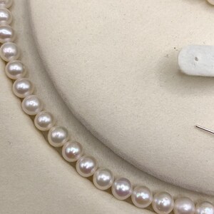 May include: A close-up of a pearl necklace. The pearls are white with a slight pinkish hue, strung together to form a delicate necklace. The background is a soft, off-white color, highlighting the necklace's elegance.