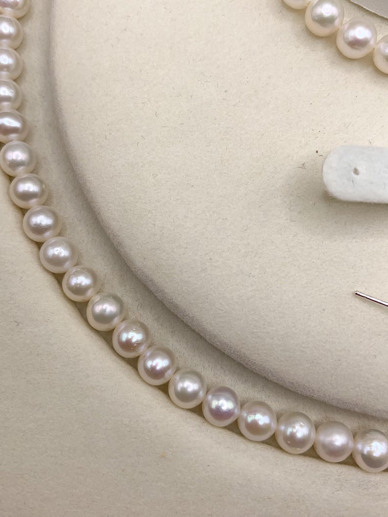 May include: A close-up of a pearl necklace. The pearls are white with a slight pinkish hue, strung together to form a delicate necklace. The background is a soft, off-white color, highlighting the necklace's elegance.