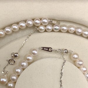 May include: A close-up view of two strands of white pearl necklaces. The necklaces have a silver clasp and extender chain. The pearls are round and have a lustrous sheen. The background is a light beige color.