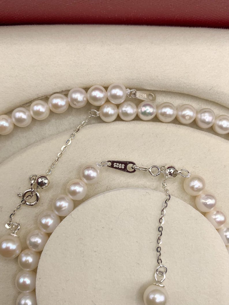 May include: A close-up view of two strands of white pearl necklaces. The necklaces have a silver clasp and extender chain. The pearls are round and have a lustrous sheen. The background is a light beige color.