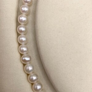 May include: A strand of luminous, round pearls. The pearls are white and evenly sized, strung together to form a necklace. The background is a soft, off-white fabric, providing a neutral backdrop for the jewelry.