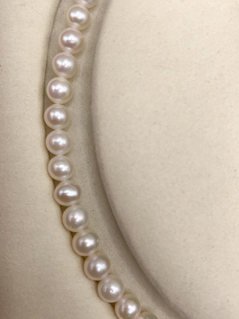 May include: A strand of luminous, round pearls. The pearls are white and evenly sized, strung together to form a necklace. The background is a soft, off-white fabric, providing a neutral backdrop for the jewelry.
