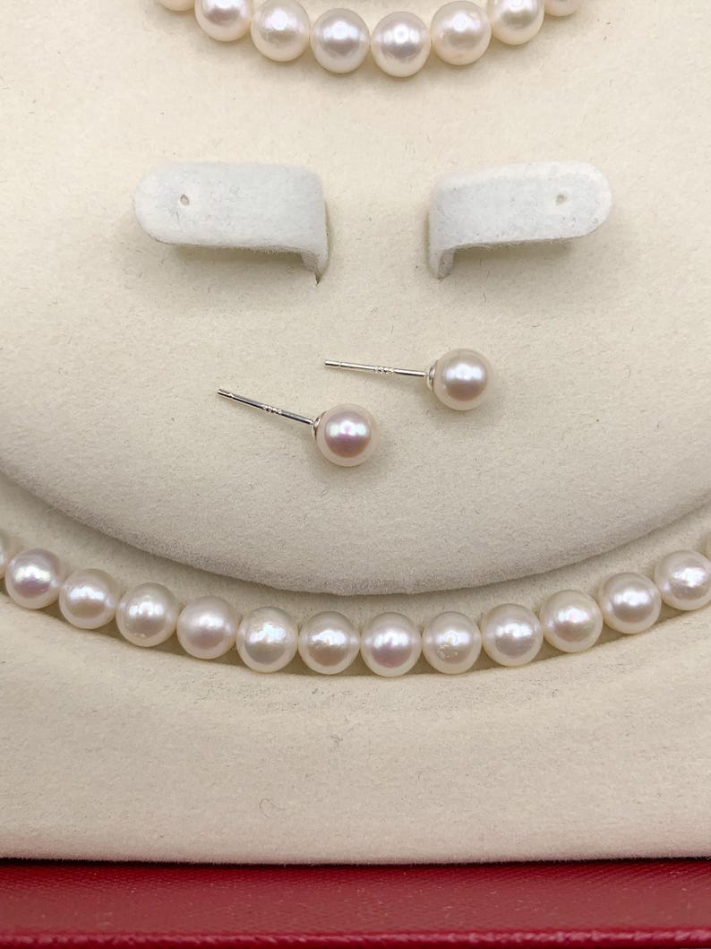 May include: A jewelry set featuring a necklace, stud earrings, and a bracelet, all made with lustrous, round, white pearls. The earrings are displayed on a white background, and the necklace and bracelet are partially visible.