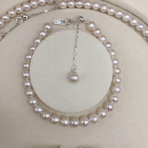 May include: A delicate pearl necklace and bracelet set. The jewelry features small, round, white pearls strung together with a silver chain. The bracelet is displayed on a white circular stand. The set includes matching pearl earrings.
