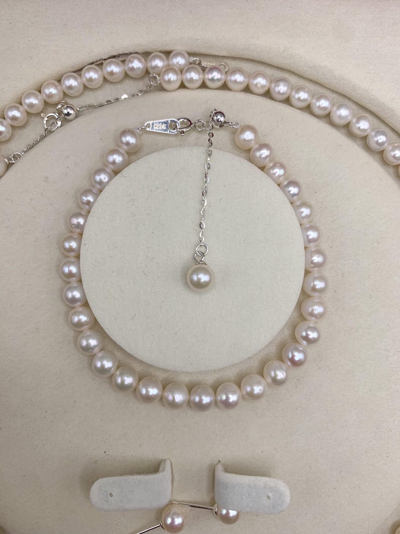 May include: A delicate pearl necklace and bracelet set. The jewelry features small, round, white pearls strung together with a silver chain. The bracelet is displayed on a white circular stand. The set includes matching pearl earrings.