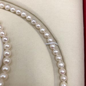 May include: A close-up of a pearl necklace and bracelet set. The pearls are white and round, strung together on a delicate string. The jewelry is displayed in a cream-colored, velvet-lined box with a red border.