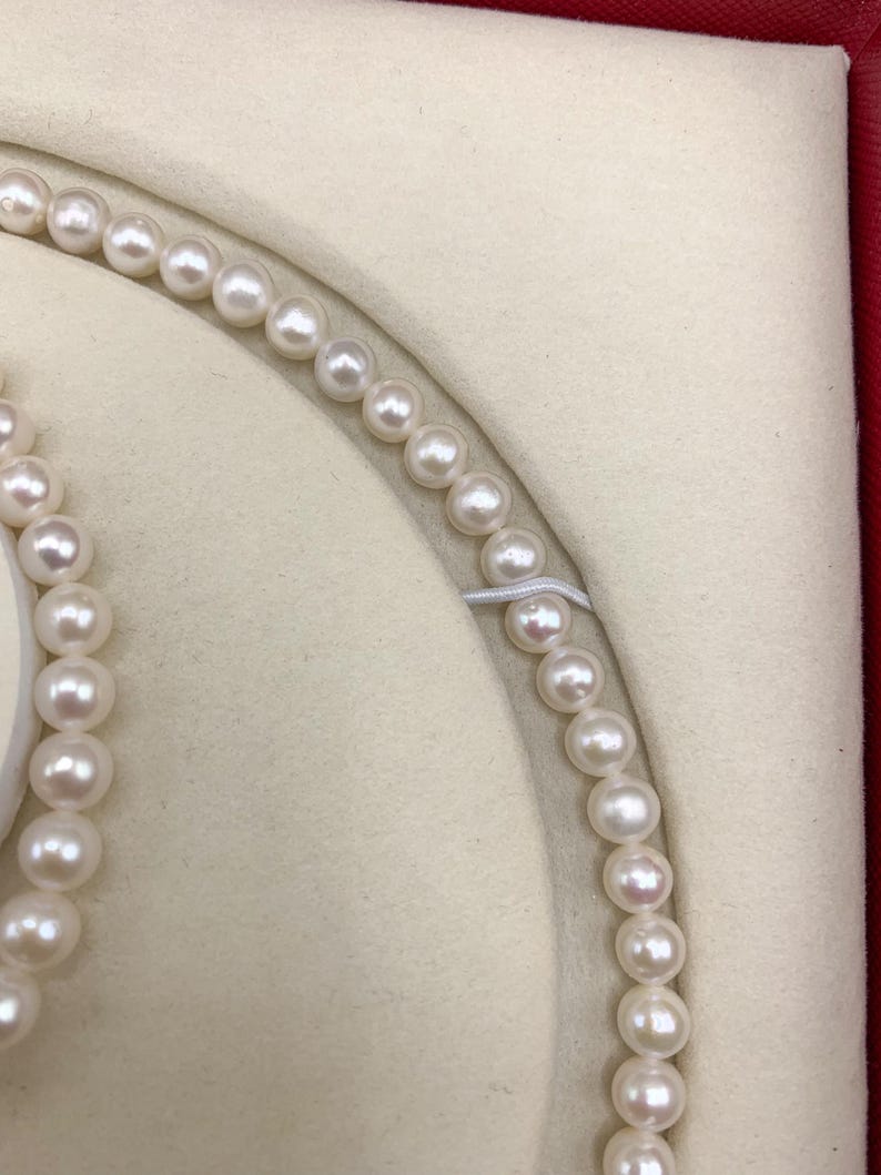 May include: A close-up of a pearl necklace and bracelet set. The pearls are white and round, strung together on a delicate string. The jewelry is displayed in a cream-colored, velvet-lined box with a red border.