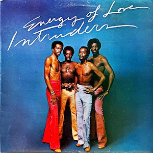 May include: Album cover featuring four shirtless men in colorful pants against a blue background. The album title "Energy of Love" and the band name "Intruders" are written in white script across the top. The men are posed, with some wearing bracelets.